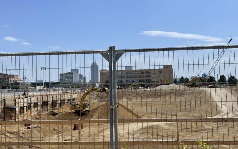 An excavator works a cleared downtown construction site behind a metal fence