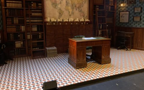 An empty stage set of a wood-paneled office with bookshelves and a desk