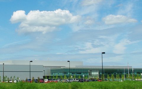 A large white industrial building and a glass office wing behind a parking lot and grass