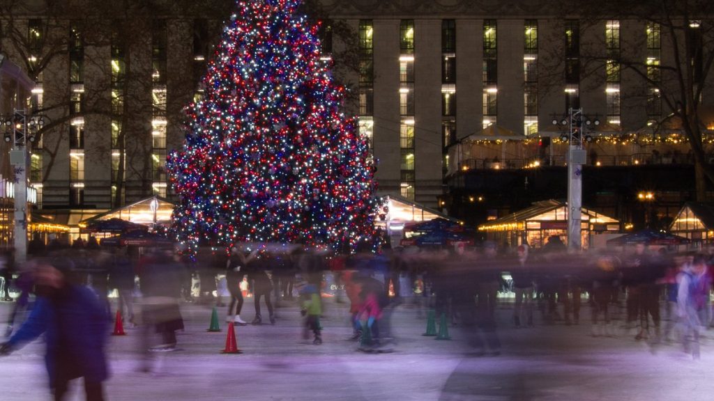 Motion-blurred skaters on an outdoor rink in front of a large lit holiday tree