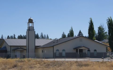 A metal-sided community building with a minaret tower behind a chain-link fence